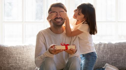 A young girl covers her father's eyes while he holds an unwrapped gift and smiles