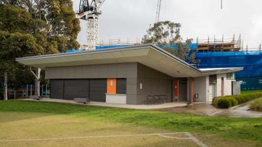 Single-storey grey and white building on the edge of a sports field, with bicycle racks and two bench seats around its outside. There is a construction crane and trees in the distance.