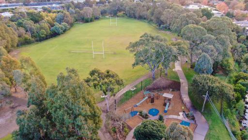 Aerial view of playground to the south and sportsground with rugby goal posts to the north. There are walking paths on each side of the playground and thick mature trees surround the area.