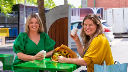 2 brightly-dressed people smile as they sip coffee at a green table