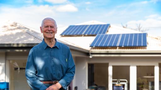An older person standing outside a residential home which has solar panels on the roof