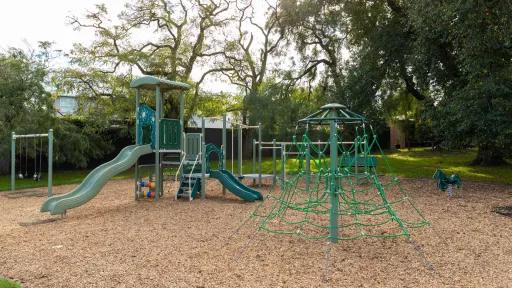 Playground with swings, rope feature and a small climbing feature with two slides. There are large trees in the background.