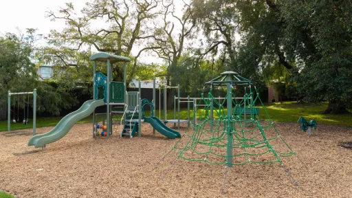 Playground with swings, rope feature and a small climbing feature with two slides. There are large trees in the background.