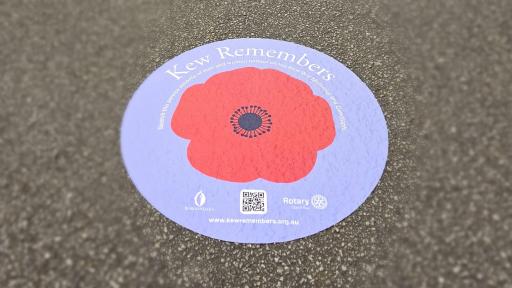 A floor decal on pavement, displaying a red poppy on a purple background with the heading 'Kew Remembers' and a qr code