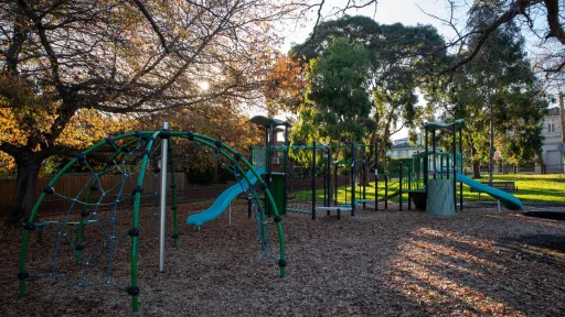 Playground in shadows with large climbing feature, two slides and a dome-shaped feature. There are tall trees and large houses in the background, with sunlight coming through.