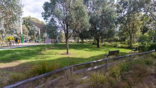 A small grass area separated from a grass shrub area by a wooden fence. The grass area has a couple of trees and wooden seats. in the background is a playground covered by a shade sail and a wooden picnic table.