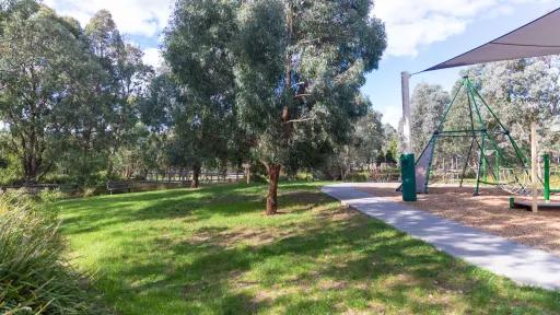 A shaded grass area with a tree in the middle. To the right is a mulched playground covered by a shadesail. A concrete path surrounds the playground.