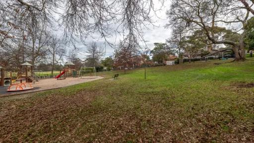 A large grass area sloping downward towards a playground with mulch surface. The playground has 2 sets of swings, 2 climbing frames. A boundary fence runs along the perimeter of the park.