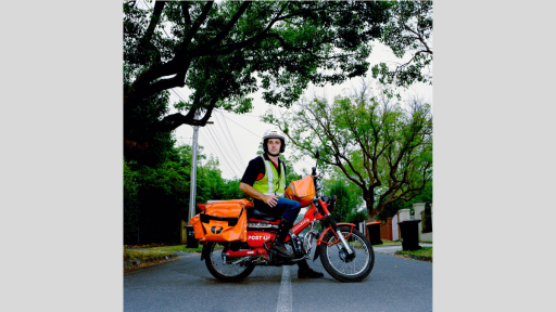 Photograph of a postman on a red Australia Post motorcycle. Orange mailbags are attached to the bike, and the postman is wearing a high-vis vest and white helmet. He is stopped in the middle of the street with one foot on the ground to steady the bike looking directly at the camera. The background shows a tree-lined street with wheely bins out for collection.