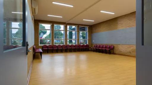A small empty room with a wooden fall. Chairs lines the back wall of the room. Large windows look out towards the tress outside.