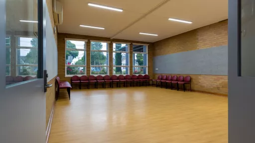A small empty room with a wooden fall. Chairs lines the back wall of the room. Large windows look out towards the tress outside. 