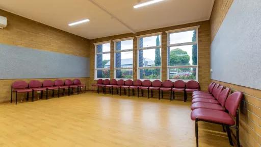 A small empty room with wooden floors. Chairs line the back walls, and large windows look out to trees outside.