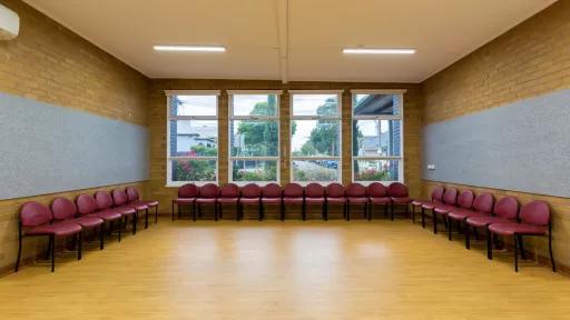A small empty room with wooden floors. Red chairs line the back walls. Large windows look out onto trees outside.