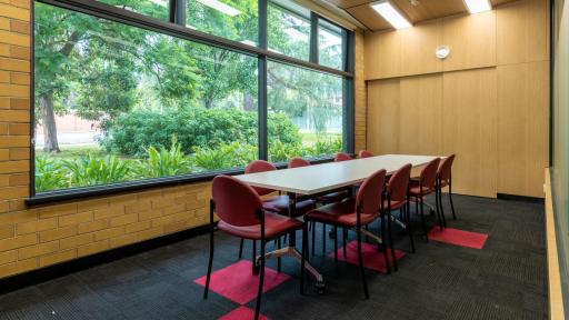 A small room with a long rectangular table surrounded by red chairs. A large window overlooks the trees outside 