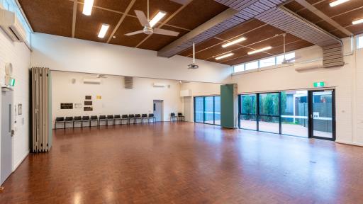 A large empty room with wooden floors. Tall windows look out to an outdoor area. Retractable wall panelling can be seen on both sides of the room. 