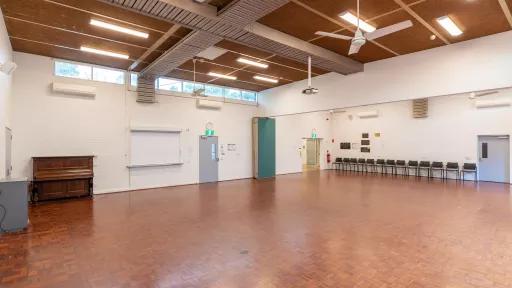 A large empty room with wooden floors. Chairs line one wall of the room,