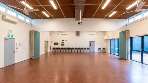 A large empty room with wooden floors. Chairs a lined up against the back wall and light is coming in from large windows on the side.