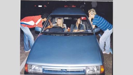Photograph of two young women sitting in a parked car speaking to two young men leaning through the front windows. The car is an old blue Ford, and a red ‘P’ plate is in the top right corner of the front windshield. The body language of all four people indicates the conversation is fun and flirtatious, and their clothing and hair styles are representative of the early 2000’s fashion trends.