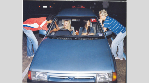 Photograph of two young women sitting in a parked car speaking to two young men leaning through the front windows. The car is an old blue Ford, and a red ‘P’ plate is in the top right corner of the front windshield. The body language of all four people indicates the conversation is fun and flirtatious, and their clothing and hair styles are representative of the early 2000’s fashion trends. 