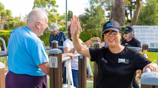 A group of people outside in a park area. 1 person wearinng reflective sunglasses is looking directly at the camera and waving