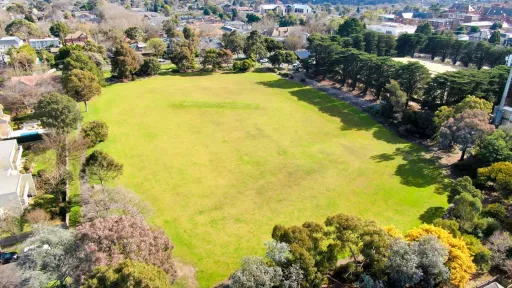 A overhead view of a large sportsfield surrounded by large trees. A cricket pitch is in the centre.