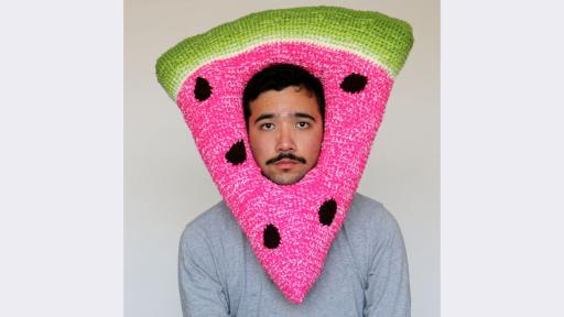 Image of a figure wearing a crocheted watermelon slice, shaped as a triangle with a hole in the middle fitted around their face. The figure has short, dark hair, a dark moustache, and a grey shirt. Their face is expressionless. The watermelon ‘hat’ is bright pink with black spots, and the top of the hat features a bright green rind.