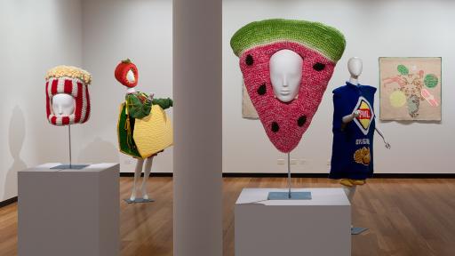 Four mannequins at an exhibition wear unique outfits: the first wears a yellow apron and a popcorn headdress, the second wears a watermelon headdress and a pink dress, the third wears a striped shirt and a popcorn box headdress and the fourth wears a colorful dress and a tomato headdress.