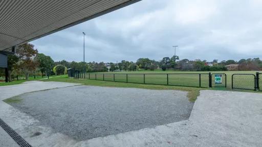 A view of the fenced sportsground. Surrounding the field is a concrete and gravel area under a roofed area.