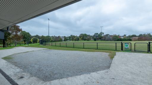 A view of the fenced sportsground. Surrounding the field is a concrete and gravel area under a roofed area. 
