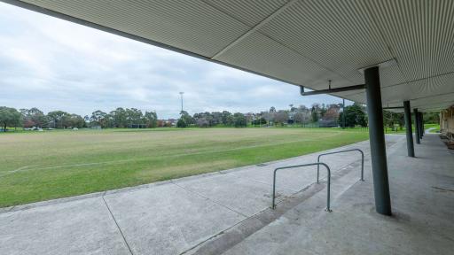 Looking out towards a green sports field from a sports grandstand . Trees and houses are visible in the background. 