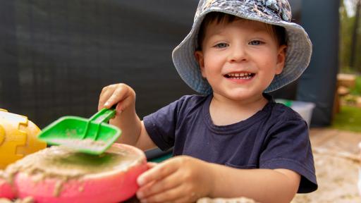 Young child in blue patterned hat plays with green spade in sandpit.