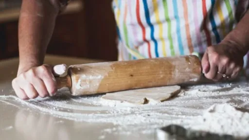 photo of a person rolling dough with a rolling pin. They are wearing a striped apron and there is lots of flour on the bench.