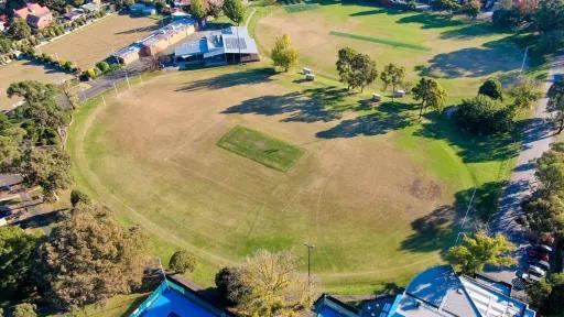 An aerial view of a green sports oval surrounded by trees. A building sits on the edge of the oval. a second sports oval sits beside it. 