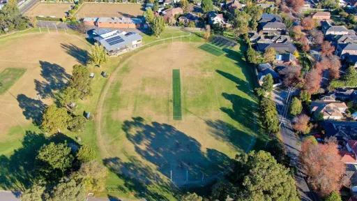 Aerial view of an oval sportsgrouind. A green cricket pitch is in the centre. The oval is surrounded by houses and trees. 