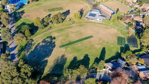 An aerial view of an oval sportsground. It has a green turf cricket pitch, and cricket nets. A second oval can be seen in the background.