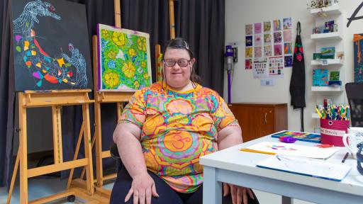 Artist Miranda Millane wearing a tshirt with a brightly coloured mandala. Behind her are 2 paintings. one has yellow flowers surrounded by green leaves with blue filling in the spaces between. The other has white hands painted on a black background. A red string is tied between 2 fingers. between the hands is a tangle of lines, with some spaces filled in with bright colours.
