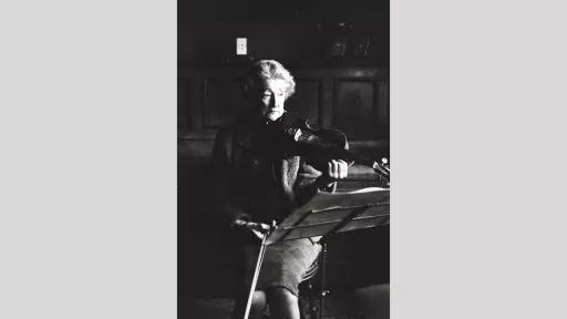 A black-and-white photograph featuring an older woman seated and playing a violin. She is dressed in a dark coat and appears to be deeply focused on her music, with her eyes partially closed and her hands positioned on the instrument. In front of her, a music stand holds sheet music. The background shows a dimly lit interior with wooden panelling and a framed picture, creating a nostalgic and intimate atmosphere.