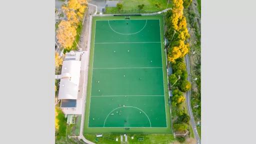 An aerial view of a rectangular sports field, presumably a hockey field, featuring a synthetic green playing surface marked with white lines. The field is surrounded by a boundary with a buffer zone of artificial turf or grass. Several small figures, likely players, are scattered across the field engaged in various activities. On the left side of the image, adjacent to the field, there is a white-roofed building, possibly a clubhouse or facility associated with the sports field.