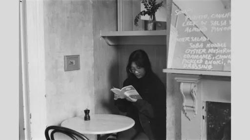A black and white photo of a woman reading a book in a cozy café. She is seated at a small table next to a wall-mounted chalkboard menu. The scene exudes a quiet, intimate atmosphere with minimalistic décor.