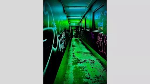 A narrow, indoor corridor with walls covered in vibrant graffiti. The corridor is bathed in a greenish light, creating a surreal and edgy atmosphere. The graffiti art features various tags and designs, adding to the urban aesthetic.