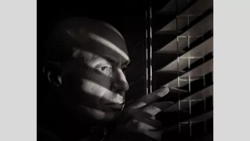 A dramatic black-and-white photograph of a man peering through partially opened blinds. His face is illuminated by slats of light filtering through the blinds, casting shadows across his features. The man appears to be intently looking outside, with one hand gently pulling down the blinds for a better view. The image conveys a sense of curiosity and tension, enhanced by the stark contrast of light and shadow. The man's bald head and intense expression are prominently featured, drawing focus to his vigilance