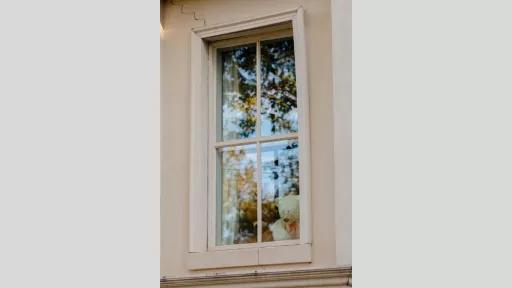Photograph of two teddy bears in a long rectangular window. The teddy on the left is brown and the one on the right is white with a red bowtie around its neck. The façade of the house is painted a light coral colour, and the outside of the window reflects the trees outside.