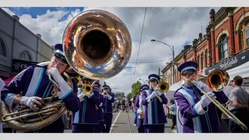 A lively street scene featuring a brass band dressed in purple uniforms with white and blue accents. The musicians are playing various brass instruments, including a sousaphone, trombones, and trumpets. The background shows a bustling street with shops and people.