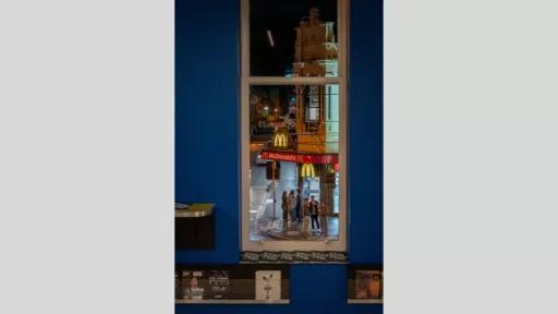 A nighttime urban scene viewed through a window. The image captures a McDonald's restaurant sign illuminated in the night, with people gathered on the street below. The scene is framed by the window, offering a glimpse of the city life outside