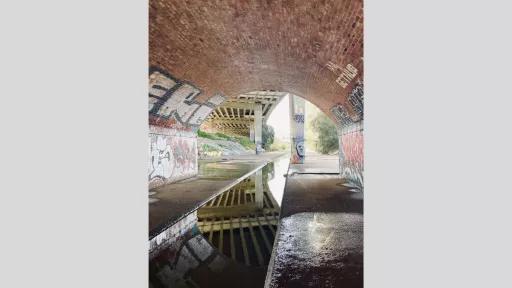 A photograph taken under a bridge with graffiti-covered walls. The scene shows a puddle reflecting the bridge's arch and the graffiti, creating a symmetrical visual effect. The background features another bridge and a path extending into the distance.