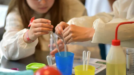 Close up view of 2 children holding pipette's in a blue cup with other science experiment items nearby.