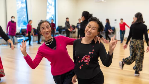 2 older chinese women smiling at the camera holding up their hands in a peace sign. Others around the room are doing stretches