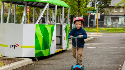 Child wearing a red helmet on a scooter rides past a small scale tram