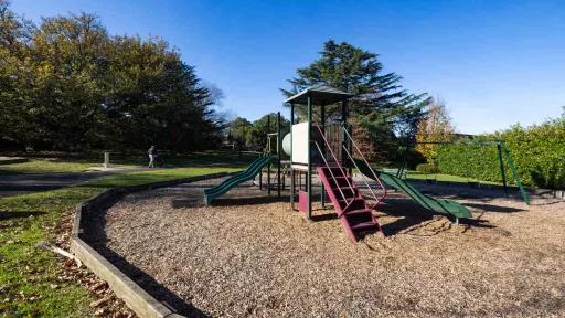 A close up of a children's playground with a tambark floor featuring 2 slides. Behind the playground is a walking path.