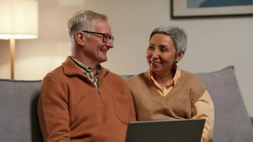 A man and woman sitting on a couch looking at each other smiling. The man has a laptop on his lap.